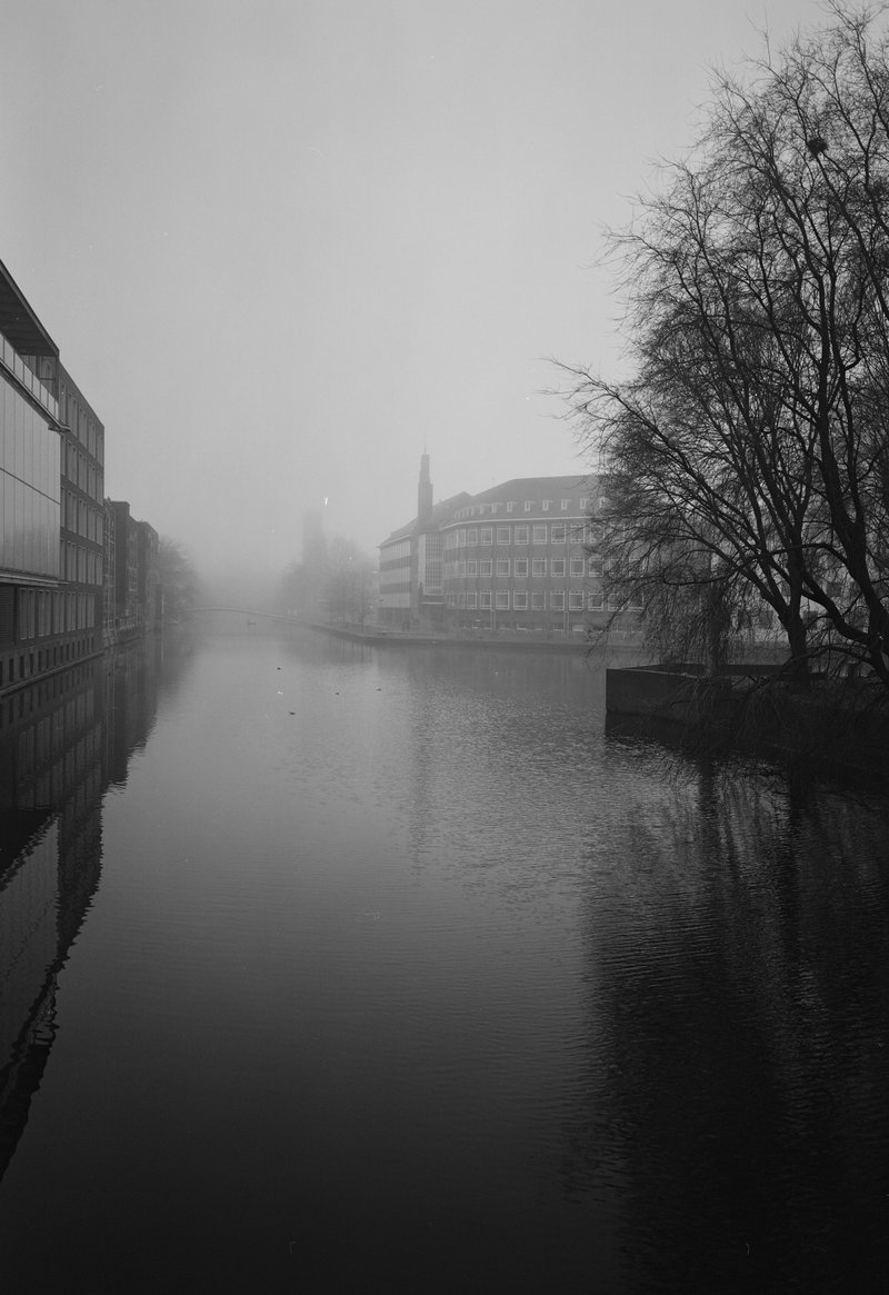 The image depicts a tranquil scene of a river flowing through a cityscape with buildings in the background, under a foggy sky. The waterway is calm, reflecting some of the overcast light above. On one side of the river, there are trees and what appears to be some greenery or park area, suggesting a peaceful urban environment.