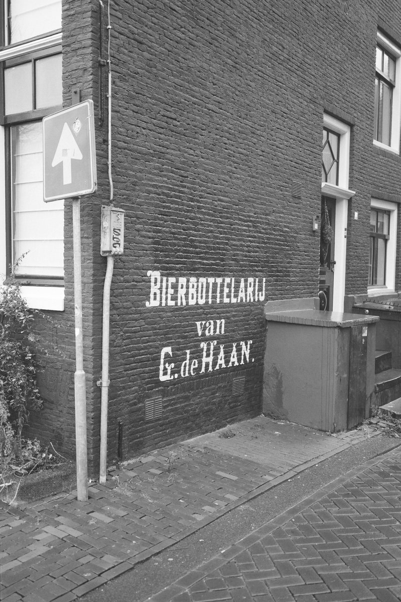  This is a black and white photo of an old brick building, likely a bar or tavern, with an advertisement that reads "KAAS en CAFE" indicating its function as a place for both food and drink. A sign says "Bierboutique" which might indicate the type of beers they offer, possibly a specialized selection. To the left of the building, there's a sign pointing towards the sea or harbor, suggesting this location is near a body of water. The overall scene appears to depict an urban area with historical architecture and a connection to maritime activities. 