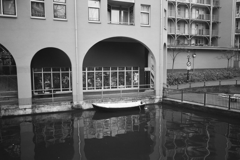  The image shows an old town scene with a boat docked near the edge of water under an archway. It seems like a rainy day as suggested by the wet street nearby. There is no text visible in the image. 