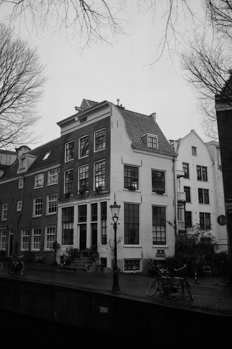  The image depicts an old European building with a distinctive row of small windows, situated on a street lined with trees. The facade of the building is adorned with ornate decorations, hinting at historical significance. The photograph has a vintage or artistic effect applied to it. 
