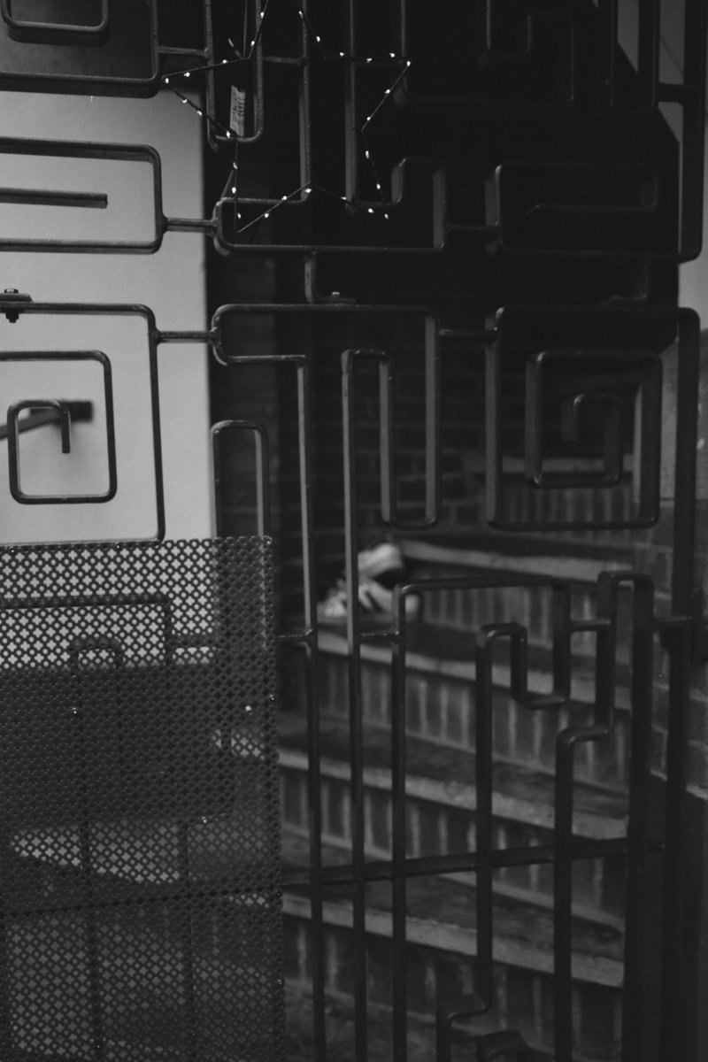  The image depicts an urban night scene with bars visible in a window. Through the bars, there's a view of a building facade. A single person is seen walking outside the barred window. There are no distinguishable features or colors. The image may evoke a feeling of solitude or confinement due to the barred window. 