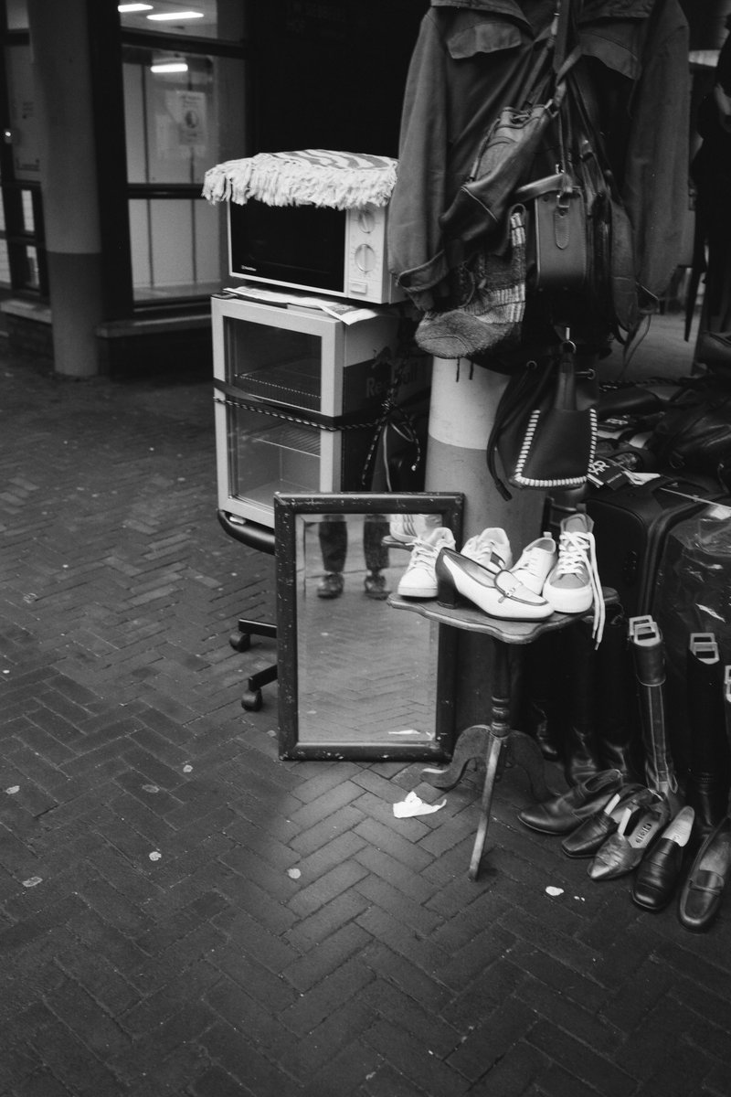  The image is a black and white photograph of an indoor scene. In the foreground, there are multiple pairs of shoes displayed on racks, with some being placed inside a box. A coat is hanging nearby. In the background, a shop window displaying items can be seen. The setting seems to be a retail space, possibly a shoe or clothing store. 