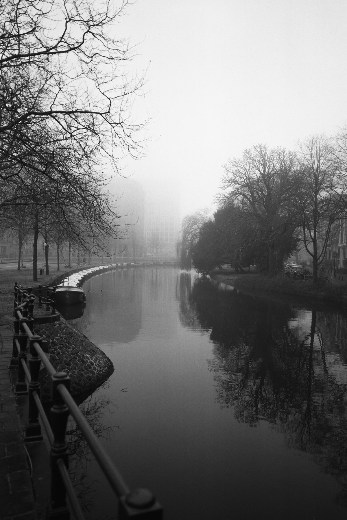  This is a black and white photo of a river or canal with trees bordering it. In the foreground, there's a fence running parallel to the waterway. The trees are leafless, suggesting it could be fall or winter. There's a bridge in the background spanning over the water. The water is calm and reflective, indicating it's likely a quiet time of day with no boats visible. A bench is situated on the side of the river, offering a place to sit and enjoy the view. There are buildings on the far bank, which provide an urban backdrop to this natural scene. 