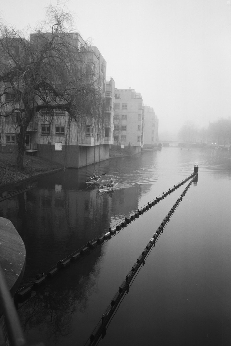  The image depicts a serene city scene with a canal running through it. On one side of the canal, there is a row of houses or apartments, while on the other, there are trees providing a natural element to the urban landscape. The sky above is overcast, suggesting an early morning or late afternoon hour when the air is usually cooler and less humid. There's no visible text or human activity, which gives the image a calm and undisturbed atmosphere. 