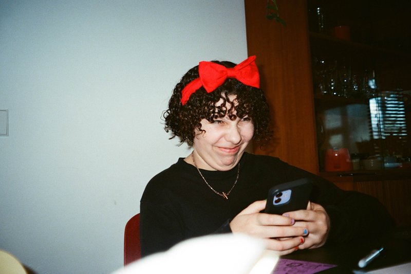 The image is a photograph featuring an individual seated with a cell phone. They are wearing a bow in their hair and have short, dark hair. The person appears to be taking a selfie as the camera is directed towards themselves. In the background, there's a glimpse of a domestic setting with a red object, possibly furniture, on the left side of the frame. The overall impression is casual and candid, likely a personal or social media post.