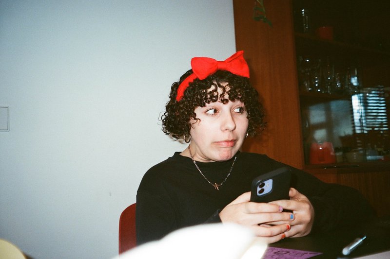 The image features a young individual sitting at a table, holding a phone. They are wearing a red bow in their hair, suggesting they might be at an event or celebration, possibly a birthday party. The person appears to be engaged with the device, possibly using it for communication or entertainment. The background is relatively simple, focusing on a wall and another individual who seems to be interacting with something else off-camera.