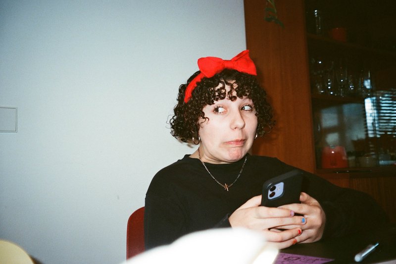 The image shows a young woman with dark hair wearing a red bow in her hair. She is seated in an indoor setting, holding a smartphone. There's a glimpse of someone or something behind her that appears blurred.