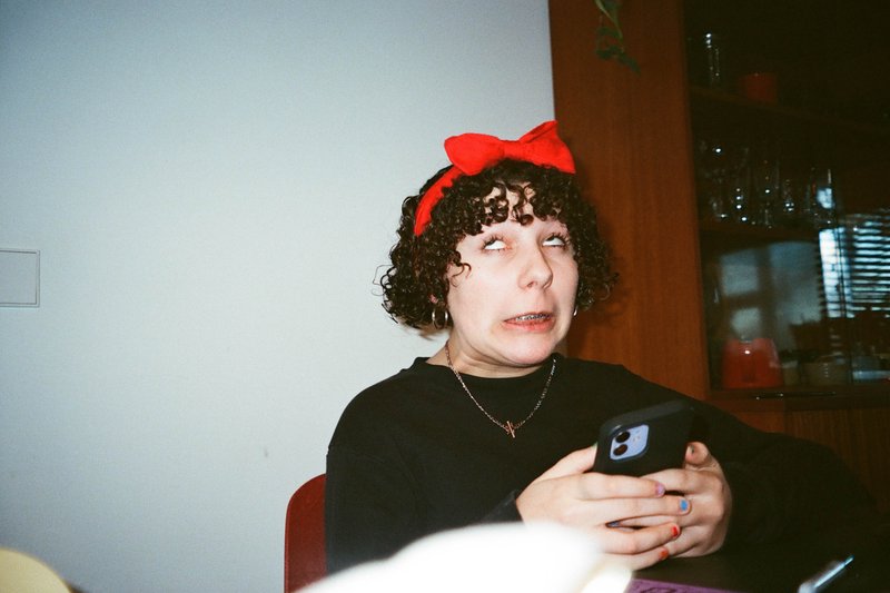 The image shows a person sitting indoors, holding a phone, with a surprised or silly expression on their face. They are wearing a red bow on their head and have short dark hair. The individual is dressed casually, in a black top, and there's a glimpse of a wall-mounted cabinet in the background. There's also a cat partially visible at the bottom of the image.