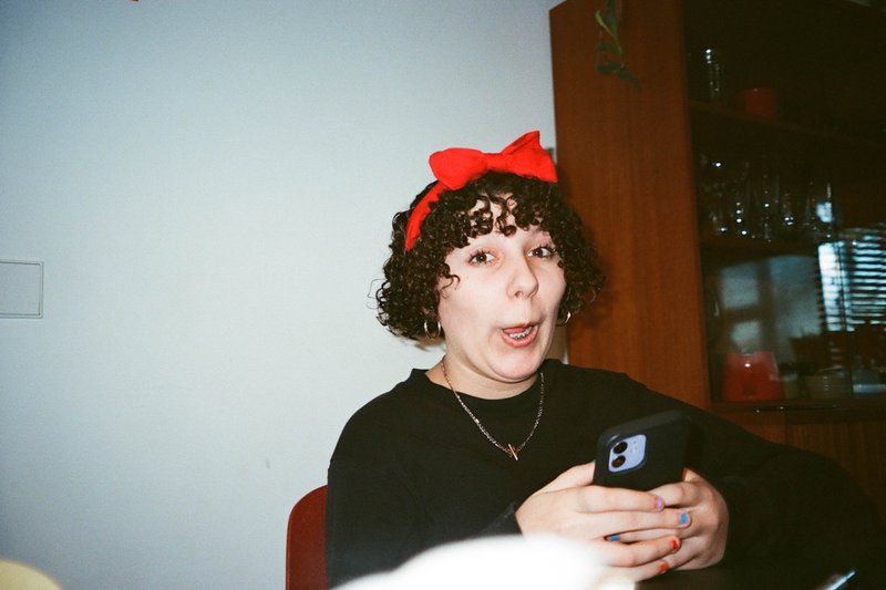 This is a color photograph featuring a young woman with a surprised or shocked expression, her hands are clasped together, and she has short brown hair. She is wearing a black top and what appears to be a red headband. There's also a playful dog in the background, adding a lively atmosphere to the scene.