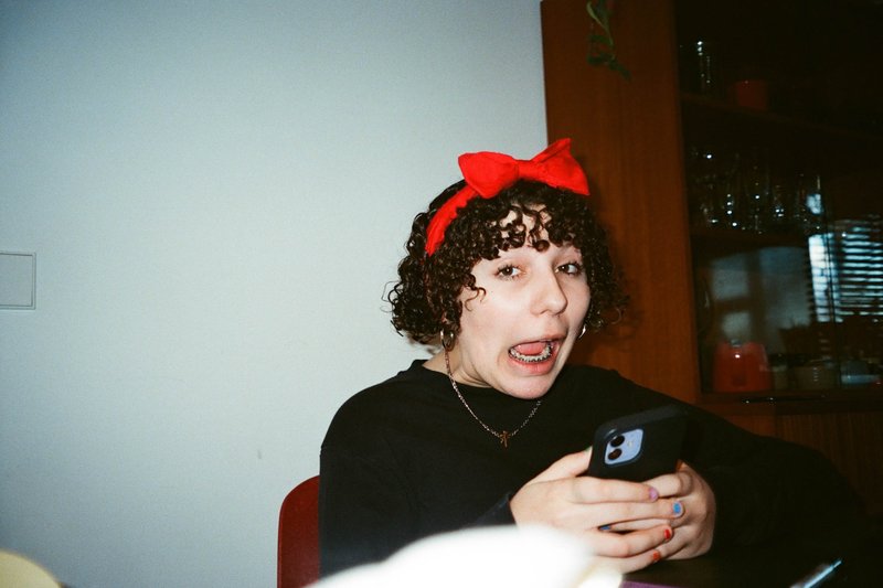 In the image, there is a young woman with curly hair who has her hair tied up. She is seated at a table, holding a mobile phone in her hand. The background of the photo features an interior setting with a red chair and a person whose face is not visible.