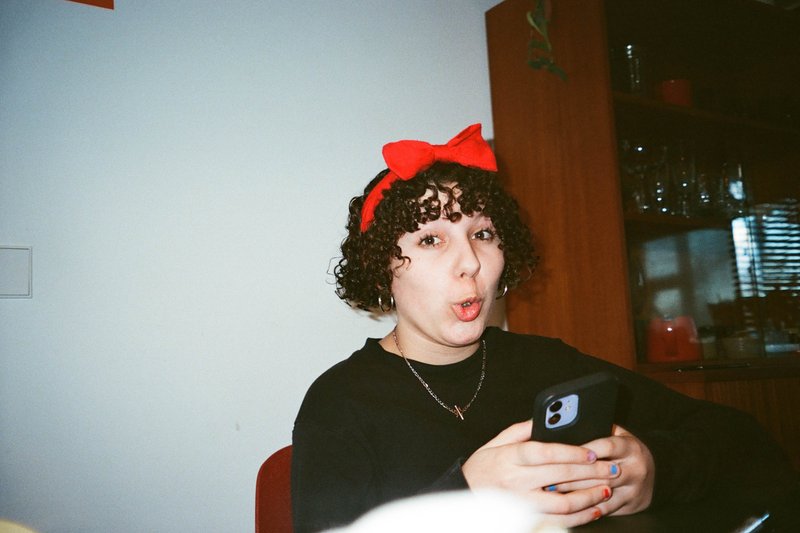 The image shows a person with short hair sitting indoors. They are wearing a black shirt and have a surprised expression on their face. Their left hand holds a smartphone, which they seem to be looking at or using. The individual is also wearing red earrings and has a red bow in their hair, suggesting a festive or celebratory occasion. The person appears to be alone in the scene, with no other individuals visible. The background features a wall with some decorations on it, and part of a chair can be seen in the corner of the image. There are no texts visible in the image.