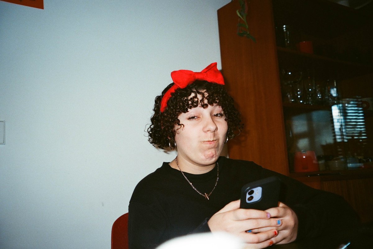  The image shows a young woman sitting comfortably in an indoor setting with a cell phone held near her face. She's wearing a playful bow, which adds a touch of whimsy to the scene. Her cheerful demeanor and casual attire suggest she's relaxed or possibly engaged in a lively conversation on her phone. 