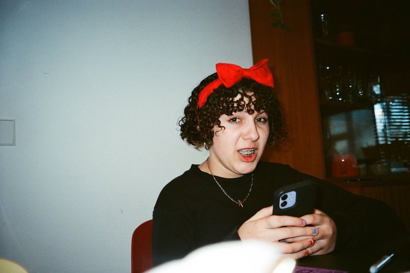 The image depicts a woman with short hair, wearing a red bow headband, seated at a table. She appears to be holding her cell phone in front of her face. There is a cat lying on the table behind her, seemingly indifferent to her actions. The background suggests an indoor setting with warm lighting and a casual, homey atmosphere.