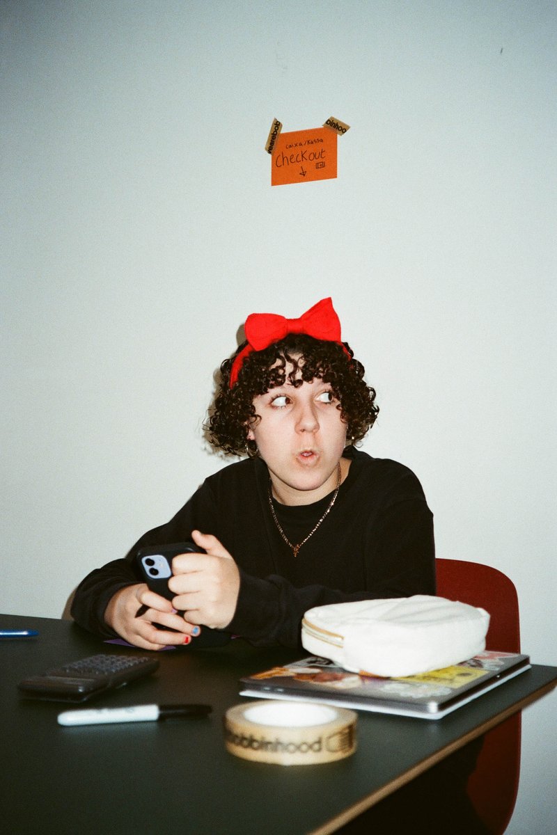 The image depicts an individual, who appears to be female, with short hair. She is wearing a black top and a red headband, sitting at a table with a cell phone on it. Behind her is a white wall with some items placed on it. Her gaze seems to be directed towards the camera.