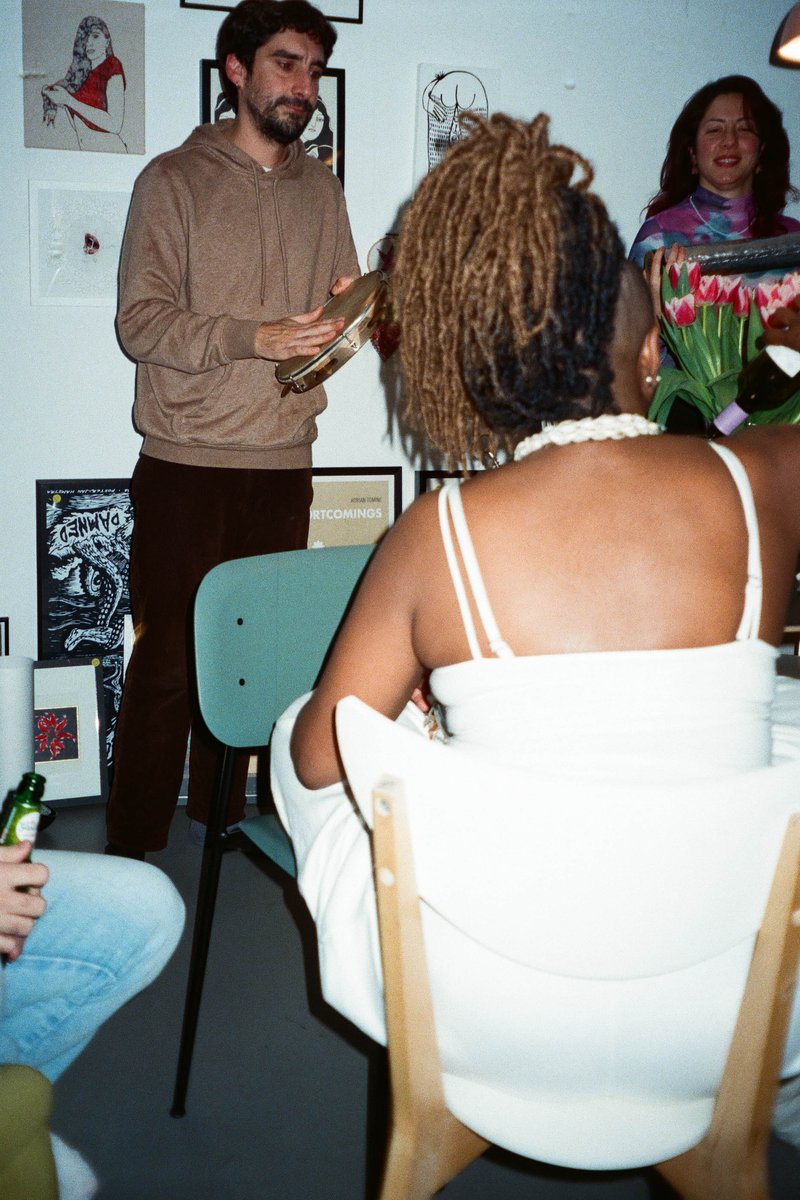 This image shows an indoor gathering, likely an art exhibition or workshop. In the foreground, a man is standing at a table with a framed item, possibly an artwork or a document, which he's holding and seems to be presenting it to a group of seated people who appear engaged in the conversation. The attendees are casually dressed, suggesting a relaxed environment. The room has an eclectic mix of decorations on the walls, including framed items and posters, contributing to an atmosphere of creativity and cultural exchange.