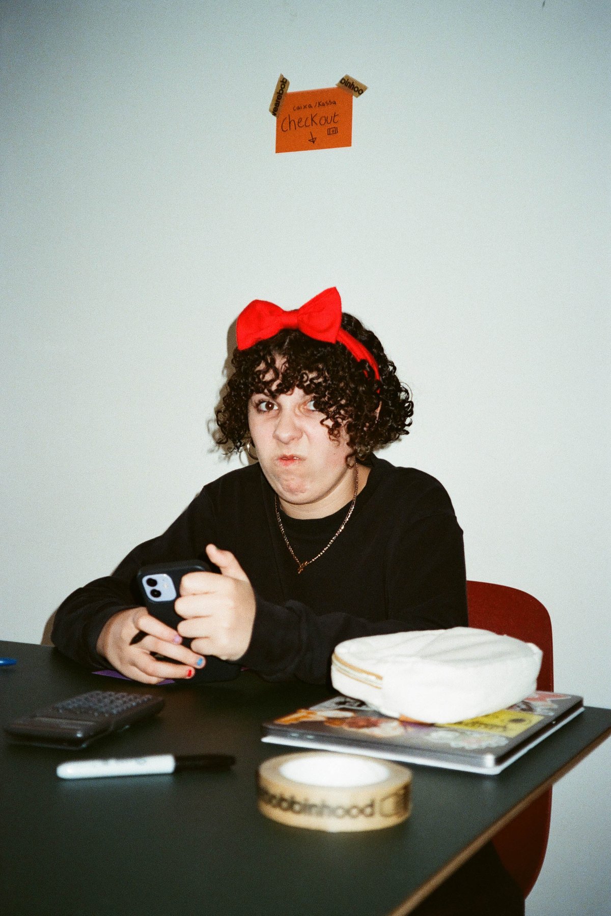  In the image, there is a woman sitting at a desk in a room with a white wall. The woman is wearing a black top and has a bow in her hair. She appears to be holding a cell phone. On the desk in front of her, there's a small jar. Behind her, there's a picture hanging on the wall. To the right, there's a book sitting on the floor. The room seems to be a casual indoor setting, possibly a living area or an office. 