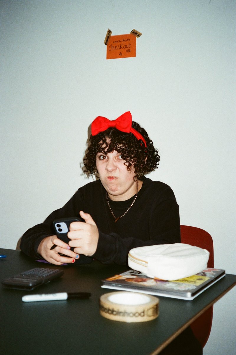 In the image, there is a woman sitting at a desk in a room with a white wall. The woman is wearing a black top and has a bow in her hair. She appears to be holding a cell phone. On the desk in front of her, there's a small jar. Behind her, there's a picture hanging on the wall. To the right, there's a book sitting on the floor. The room seems to be a casual indoor setting, possibly a living area or an office.