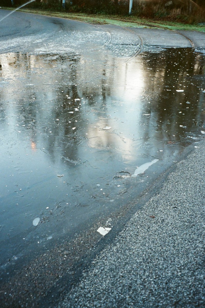 The image shows an outdoor scene with a large puddle of water occupying the foreground. To the right of this water body is a road that appears wet and reflective. In the background, there's a green landscape and trees visible under a blue sky.