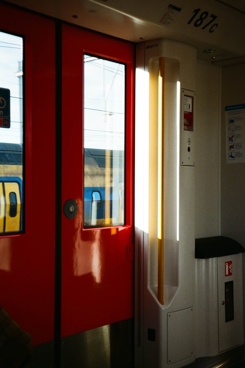 The image depicts a scene inside a vehicle, possibly a train or bus, characterized by its red doors, blue seats, and white and yellow lettering that reads "168." The sun is streaming through the windows, creating a warm, inviting atmosphere.