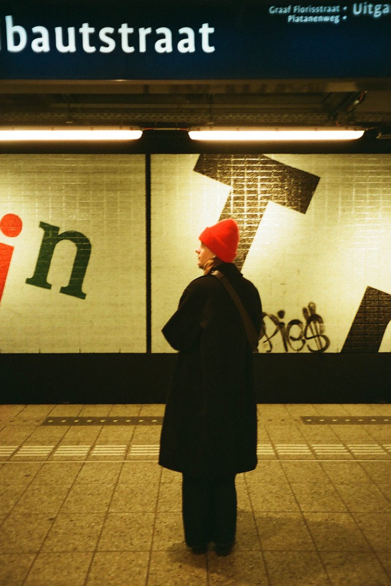 The image captures an urban night scene at a train station. The main subject is a person standing on a platform, looking out at the sign for Baustraat. The platform has advertising boards, and there's a building with a neon sign in the background.