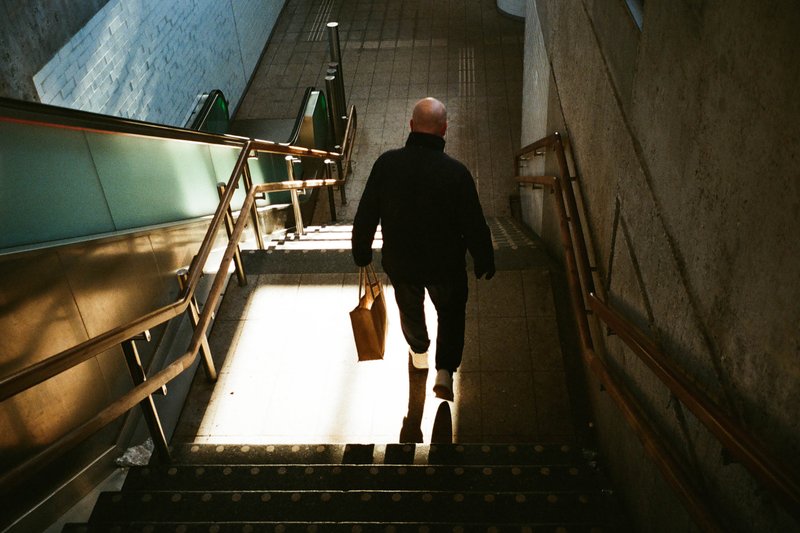 In the image, there is a man walking up a stairwell with steps visible around him. The stairwell has a high ceiling and the light coming through suggests it might be an interior space during daylight hours. There is a bag on the floor behind him. The photo appears to be taken from the same level as the stairs, giving the viewer an upward perspective.
