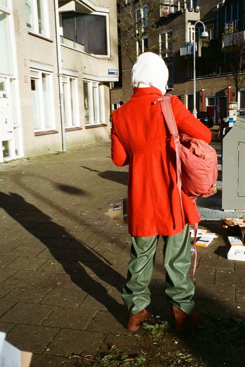 In the image, there is a person standing on a sidewalk next to a building. The individual is holding a bag and appears to be waiting or taking a break from something. They are dressed in red clothing and are wearing a white hat. The setting suggests an urban environment with houses and trees in the background.
