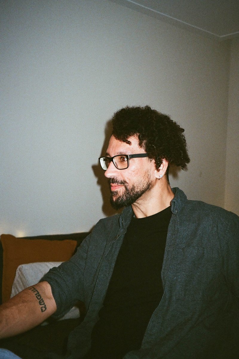 The image depicts an individual sitting in front of a white wall. This person is wearing glasses and has short hair. They are also sporting a beard and mustache. In their hand, they hold a cup which appears to contain some liquid. Behind them, a window with blinds can be seen.