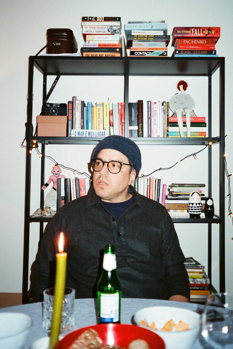 In this black and white photograph, a man is seated at a dining table which is adorned with a few books and a lit candle. The room features a festive atmosphere with Christmas decorations visible. Behind the man stands a bookshelf filled with various items, suggesting a homey setting.