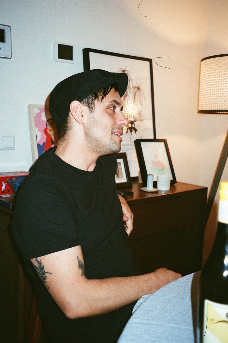 This is an image of a man sitting at a dining table in what appears to be a cozy indoor setting. He's wearing a black hat and a dark t-shirt, looking off to the side with a slight smile on his face. The background features decorative elements such as framed pictures, a light fixture, and part of another person visible through the foreground.