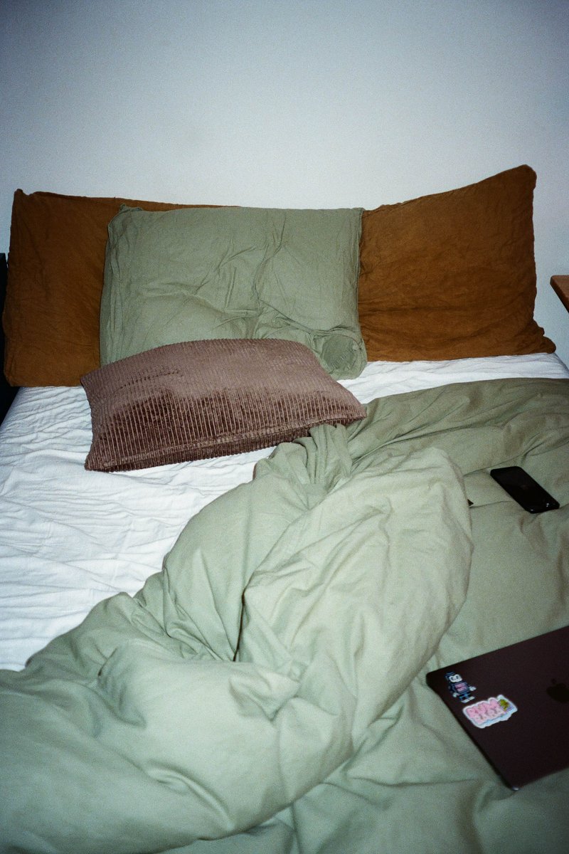 The image shows an unmade bed with rumpled sheets in shades of green and brown. There is a single pillow at the head of the bed, adorned with a dark colored case. Atop the bed, there are two folded blankets. To the right of the bed, a small black book is placed on a surface that appears to be a nightstand or table. The photograph has a grainy quality and is taken from an elevated angle looking down onto the bed.