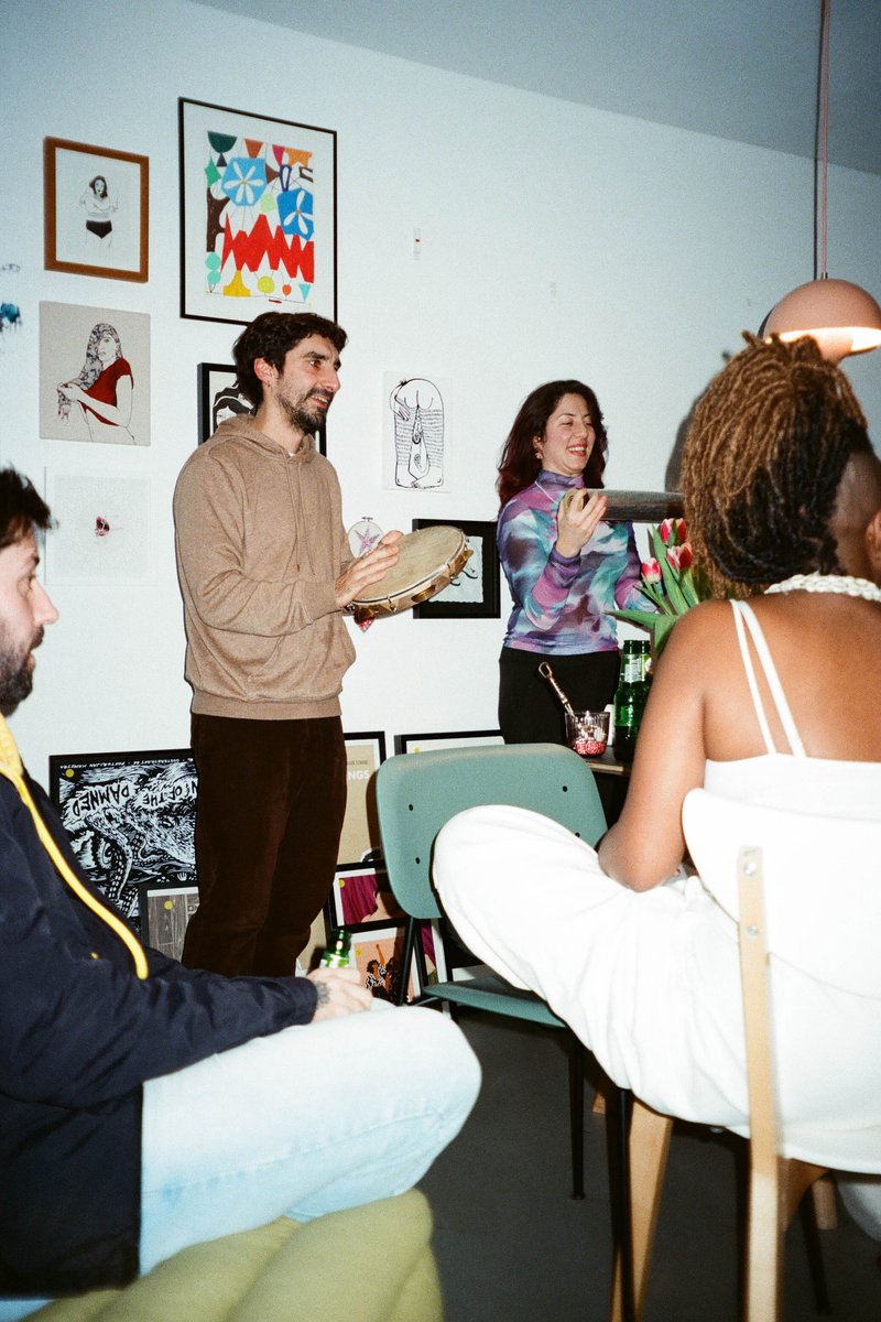 The image depicts an indoor setting where two individuals are engaged in what appears to be a conversation or presentation. One of them is holding up a framed picture, drawing attention towards it. There are other people present in the background, suggesting a casual gathering or event. The room has a warm atmosphere with paintings and artwork on the walls, providing a creative backdrop for the interaction. The lighting appears to be natural, creating soft shadows and highlighting the textures within the space.