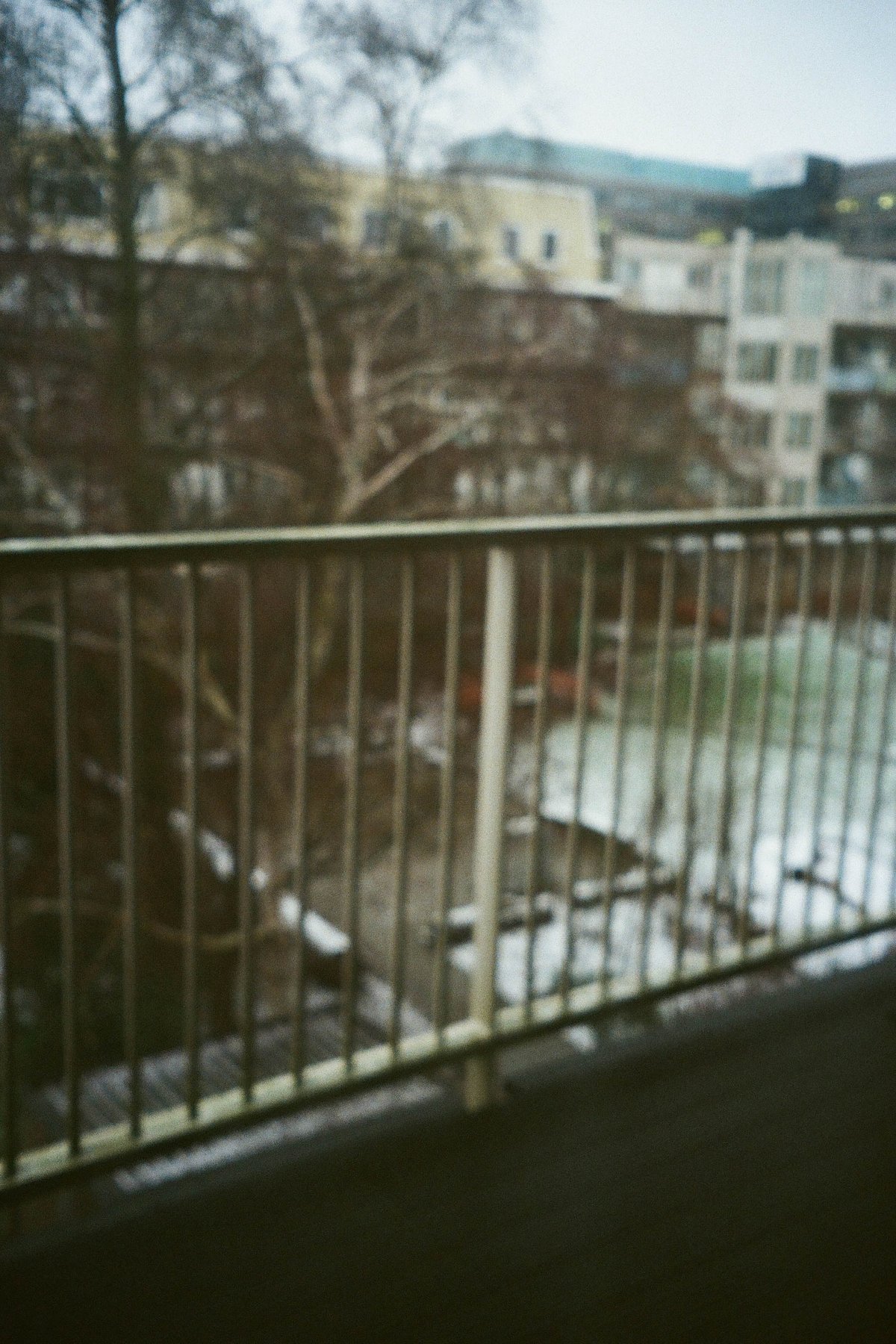  The image shows an urban scene featuring a bridge with a railing. The bridge overlooks a building and a park with trees and grass. In the foreground, there is a walkway with a metal barrier, and a portion of the sky can be seen above. The photo appears to have been taken at night or in low light conditions, as indicated by the underexposed nature of the image. 