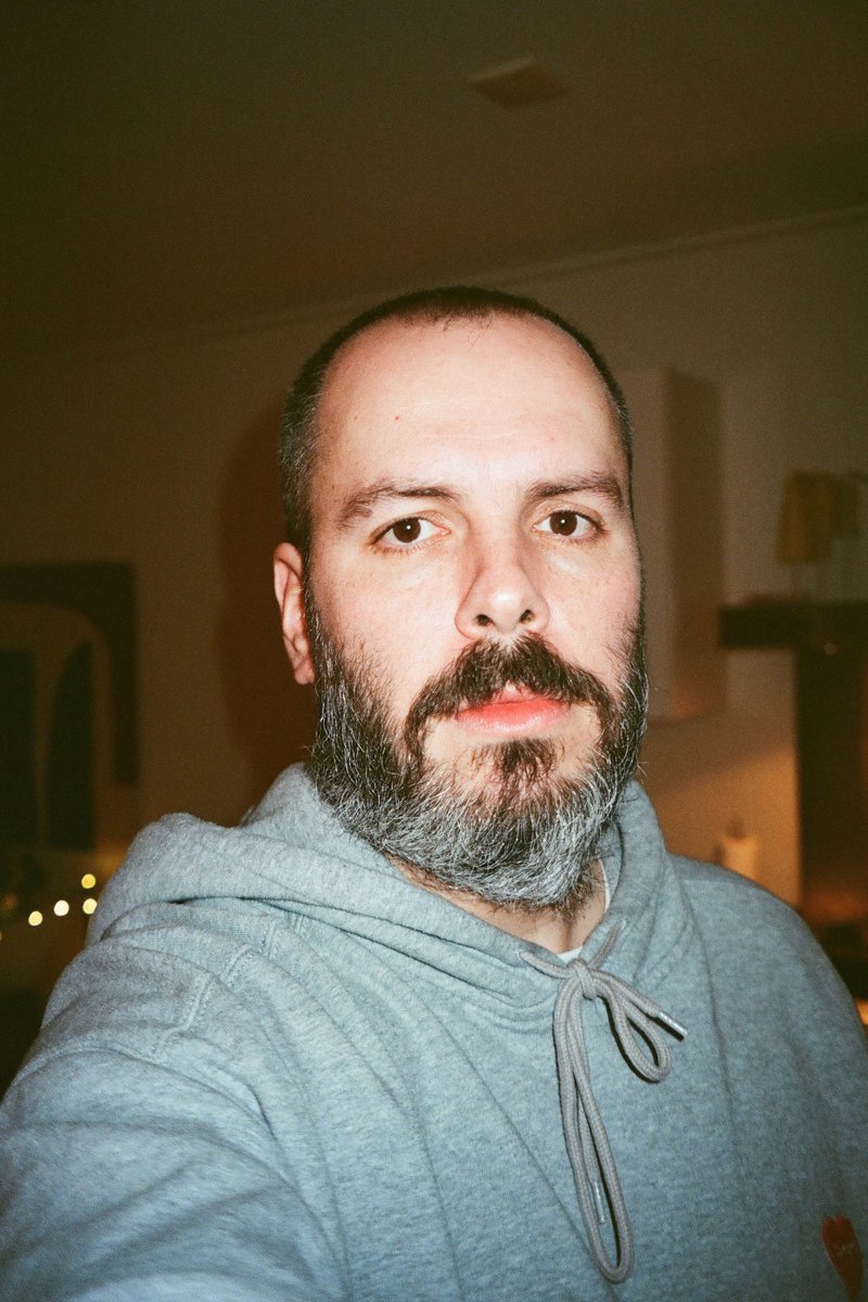 The image shows a bearded man taking a selfie. He is standing indoors with a festive atmosphere indicated by Christmas lights visible in the background.