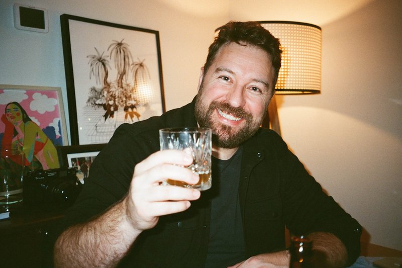 The image portrays an individual at a table, holding up a glass and smiling. They appear to be engaged in a social setting, possibly celebrating or enjoying an event. The person has dark hair and is wearing a black jacket. Their facial expression and gesture suggest a positive emotion, such as happiness or pride.