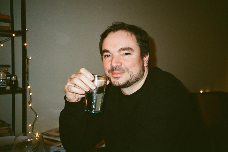 The image depicts a man seated at a dining table, holding a glass of water in his hand. He is looking directly at the camera with a relaxed expression. The background shows a dimly lit room with Christmas decorations visible. There are no other individuals or objects of significance in the image.