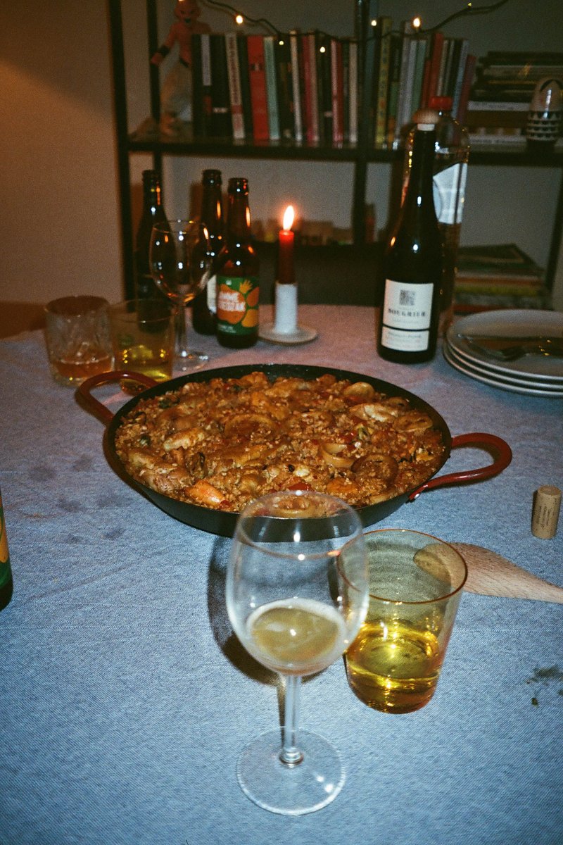 The image depicts an indoor scene where a table is set up with food and drinks. On the table, there's a tray of food, which appears to be lasagna or a similar dish, accompanied by some wine glasses and bottles. There are also some dining accessories visible, such as forks, spoons, and bowls. The background shows a room with books and decorative items, creating a cozy atmosphere.