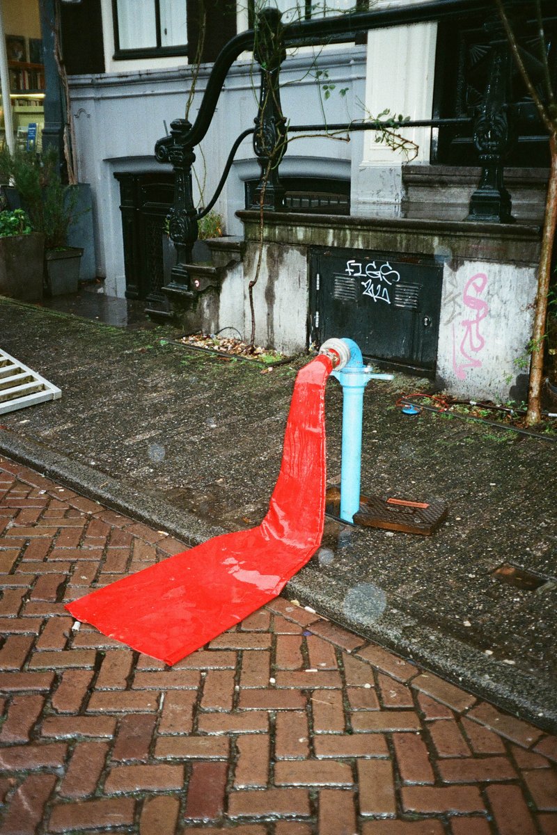 The image features a bright red ribbon or banner lying horizontally across a cobblestone street. There is a puddle beside it, indicating recent rainfall. On the right side of the frame, there is a tall blue pole standing out against the backdrop. Behind the ribbon, there's a building with a door and what appears to be a balcony or porch area. The overall scene suggests an urban setting with a touch of color added by the red ribbon.