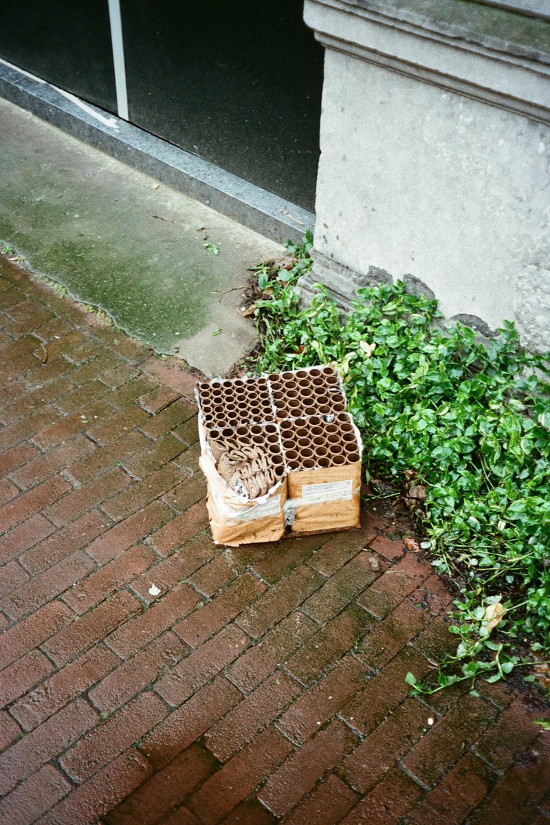 The image shows a scene of an outdoor setting with a focus on objects in front of a building. There are two boxes prominently placed on a brick pavement. One box is larger and appears to be cardboard, while the other is smaller and seems to be made of wood. The larger box has some visible text, but it's too small to read clearly. In the background, there is a black gate or fence, and part of the building can be seen as well. The ground looks wet, suggesting that it might have rained recently, which could affect the condition of the objects on the pavement.