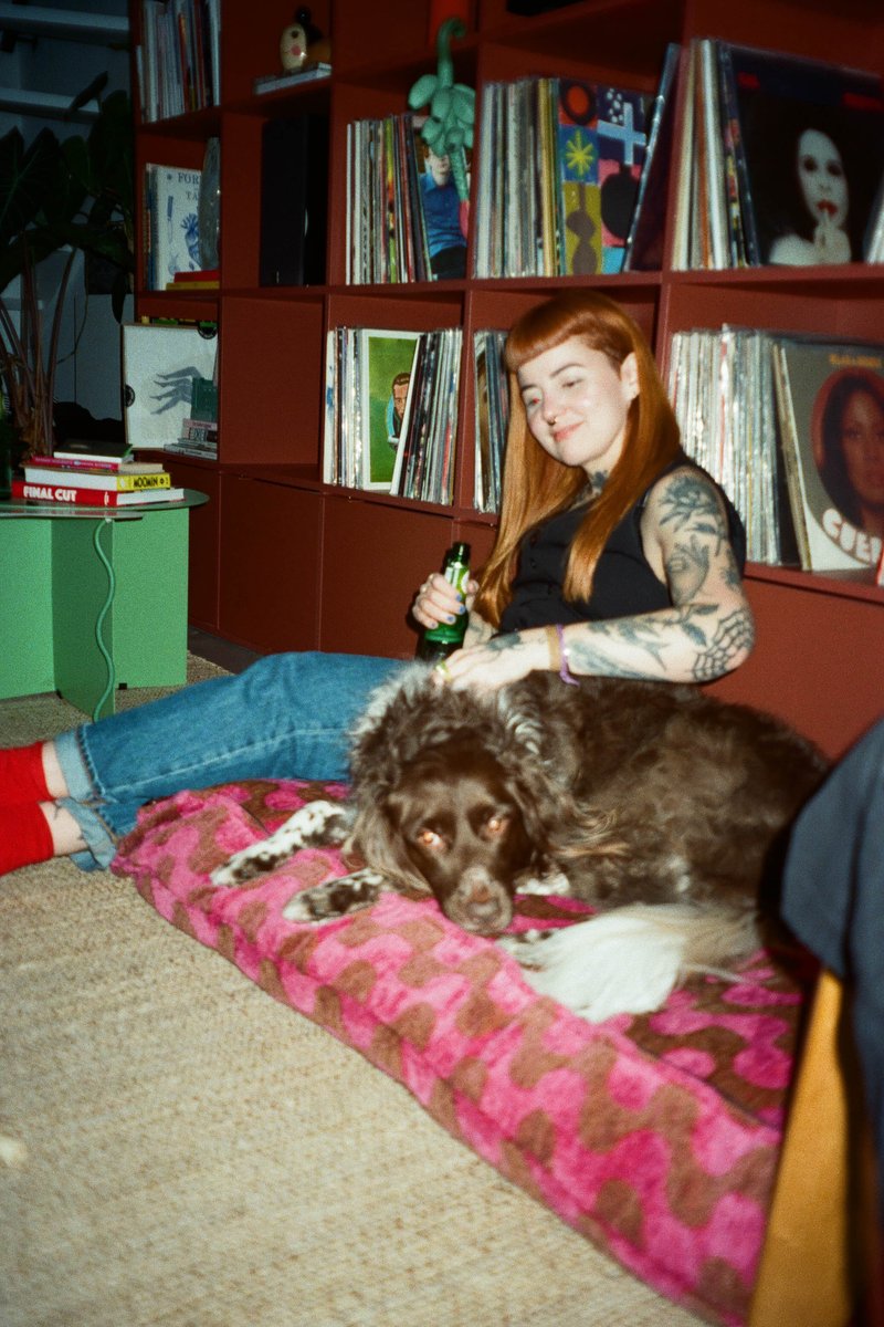 This is an indoor photo featuring a woman sitting on a cushioned floor, accompanied by her dog. She has tattoos, is wearing casual clothing, and appears relaxed with her pet. There are several books visible in the background, suggesting a home environment that includes reading or study materials. The scene conveys a sense of comfort and companionship between the human and the animal.