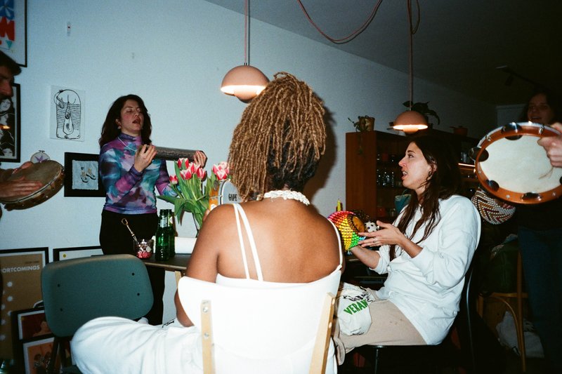 The image depicts an indoor social gathering. It features several individuals seated around what appears to be a dining table, engaged in conversation or possibly playing a game with cards. The room has warm lighting, and the people are casually dressed, suggesting a relaxed, informal setting. The atmosphere seems friendly and convivial.