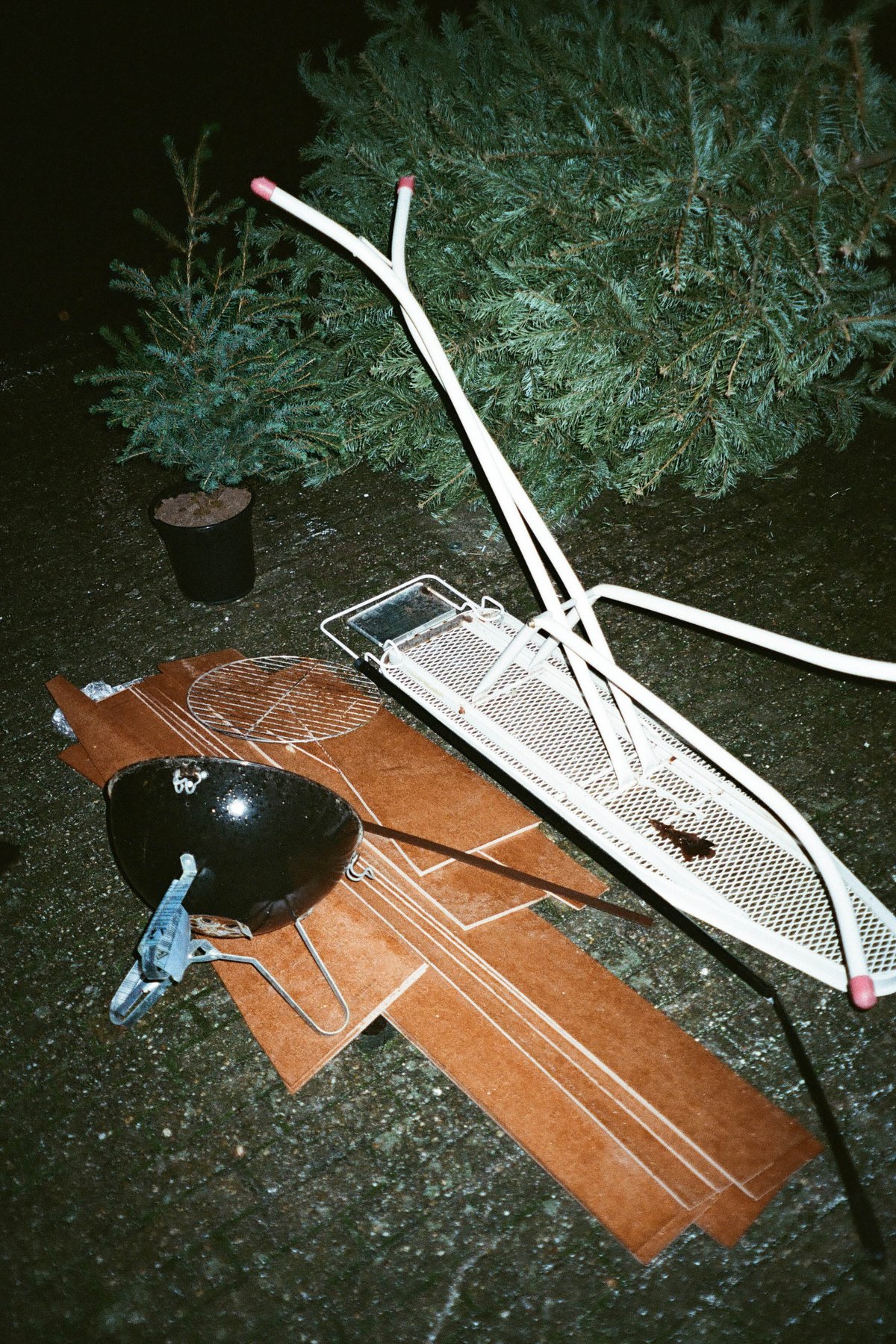  The image presents a nighttime outdoor scene featuring various objects scattered around. There's a small wooden bridge with no visible support structure lying horizontally in the foreground. Nearby, on a patch of grass, there are some broken tree branches and what appears to be a small amount of bird poop. In the background, there is a coniferous tree with its branches extending outwards, illuminated by the light coming from an unseen source. The color palette is muted, with grayscale tones dominating the image due to the nighttime setting. 