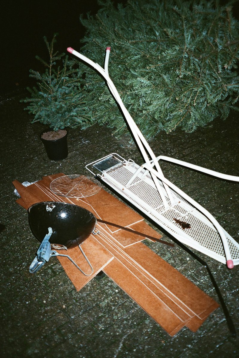 The image presents a nighttime outdoor scene featuring various objects scattered around. There's a small wooden bridge with no visible support structure lying horizontally in the foreground. Nearby, on a patch of grass, there are some broken tree branches and what appears to be a small amount of bird poop. In the background, there is a coniferous tree with its branches extending outwards, illuminated by the light coming from an unseen source. The color palette is muted, with grayscale tones dominating the image due to the nighttime setting.