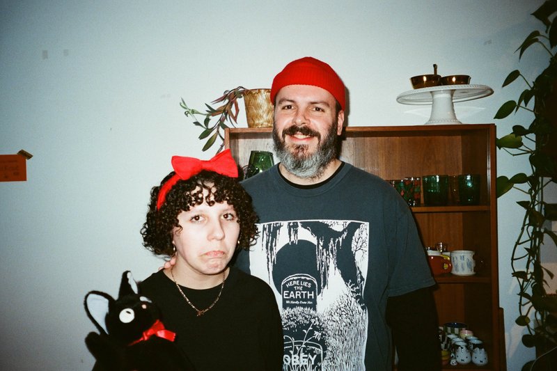 The image captures a moment of companionship between two people posing for a photo in what appears to be a cozy room. The individual on the left is standing next to a small table adorned with various items, while the person on the right is holding a stuffed animal that seems to be a cat, given its distinctive pointed ears. Both individuals are smiling and looking directly at the camera, creating a warm and inviting atmosphere.