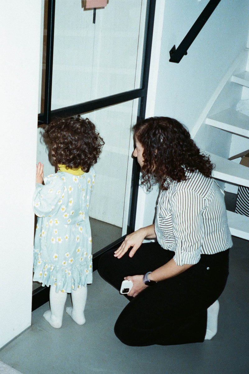 The image shows an indoor scene with a woman and a child standing together near a staircase. The woman appears to be interacting with the child, possibly showing something or addressing them. Both individuals are dressed in casual attire. The background reveals a domestic setting with a glimpse of the interior of a house. There is no visible text in the image to provide additional context.