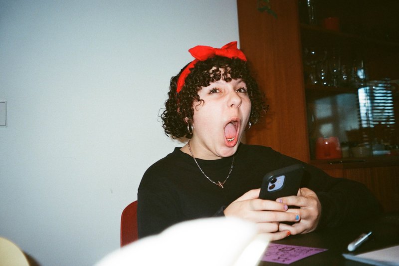 The image captures a young woman with curly hair wearing a red bow, her mouth is wide open as if she's expressing surprise or excitement. She's holding a phone in her hand and is seated at a dining table. In the background, there's a blurred figure of another person who appears to be looking at something off-camera with an expression that could be interpreted as surprise or amusement. The setting seems to be indoors, with the woman on the left side of the image and the blurred person on the right. There are no distinctive colors or specific objects that can be confidently described.