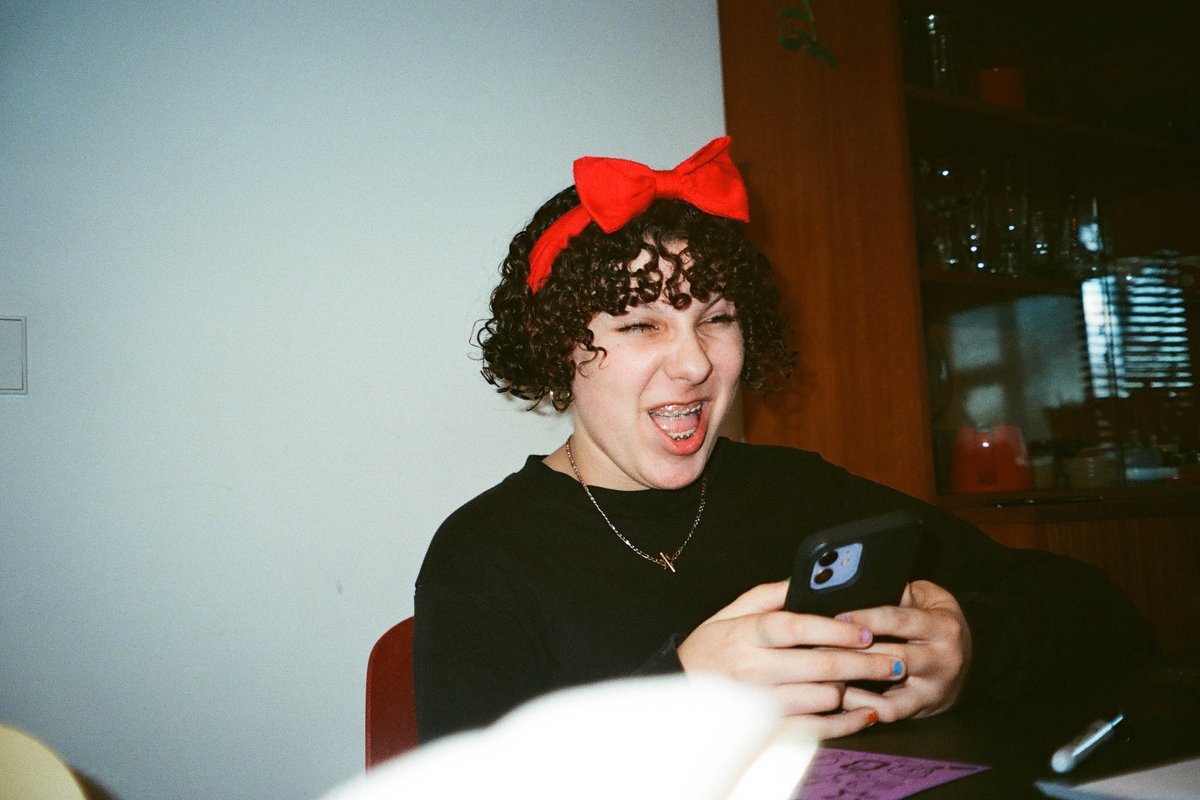  The image is a black and white photo of a person with short hair wearing a red bow on top of their head. They are smiling and looking at a phone that they're holding. There appears to be another person in the background, but their details are not as clear due to the angle and distance. 