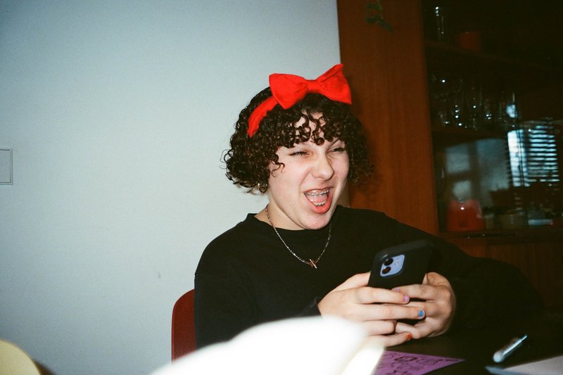 The image is a black and white photo of a person with short hair wearing a red bow on top of their head. They are smiling and looking at a phone that they're holding. There appears to be another person in the background, but their details are not as clear due to the angle and distance.