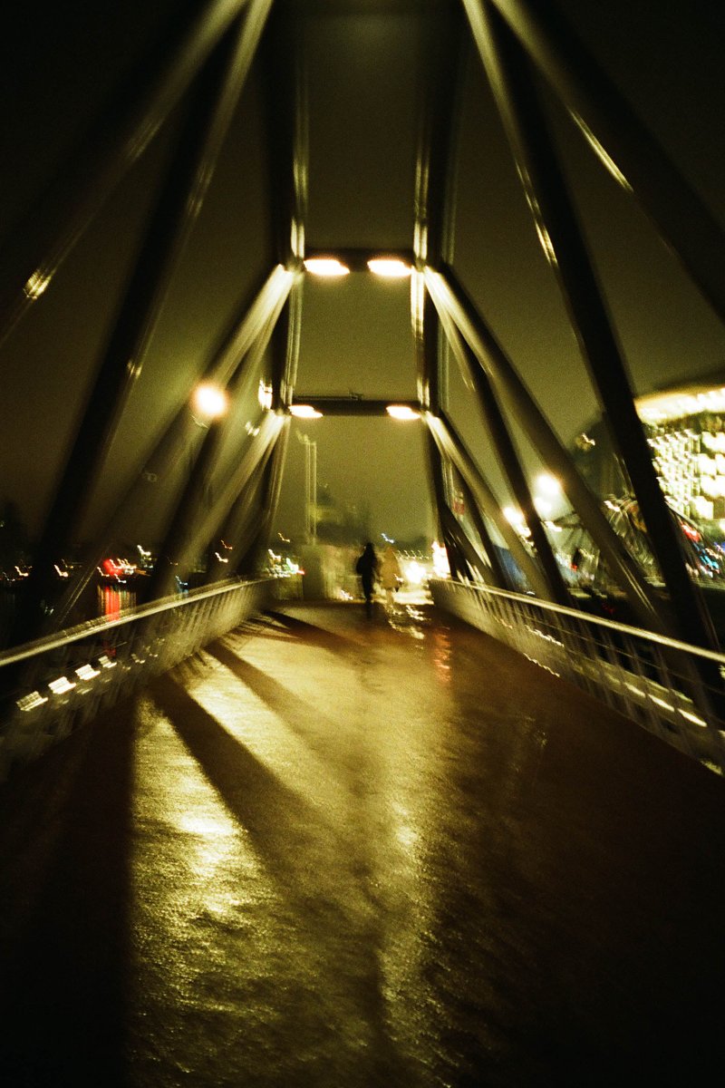 The image is of a nighttime scene featuring a dark bridge with illuminated street lights casting shadows below it. There are two people visible, walking along the bridge. The perspective is from someone standing on the ground looking up at the bridge. The bridge has a black railing and appears to be made of metal. The sky is very dark, suggesting that it is nighttime.