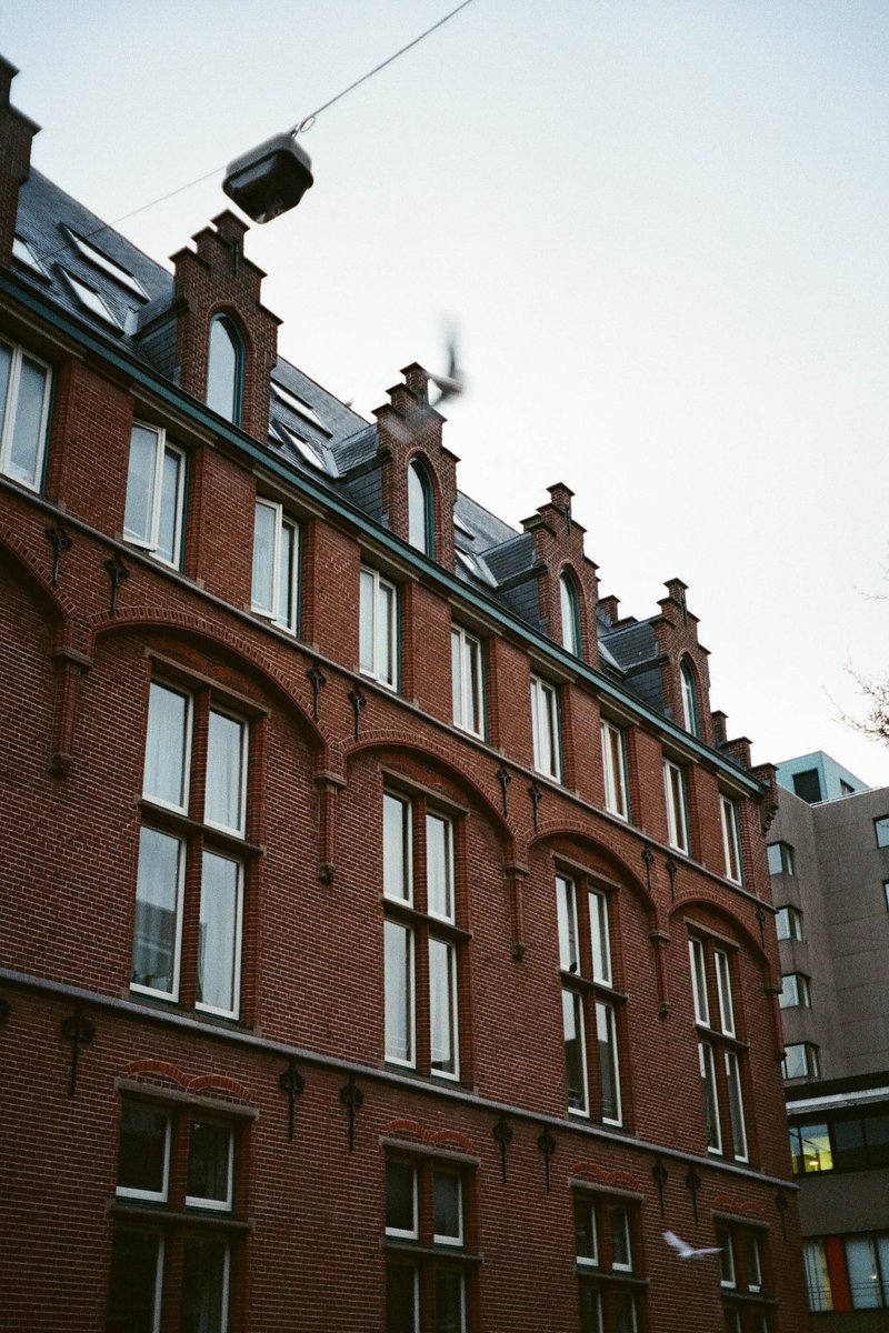 The image depicts a tall building with a distinct red brick facade, featuring multiple arched windows and gables. It's a cloudy day as suggested by the overcast sky visible above the building. There are no people or moving vehicles within the frame, suggesting a peaceful, quiet neighborhood.