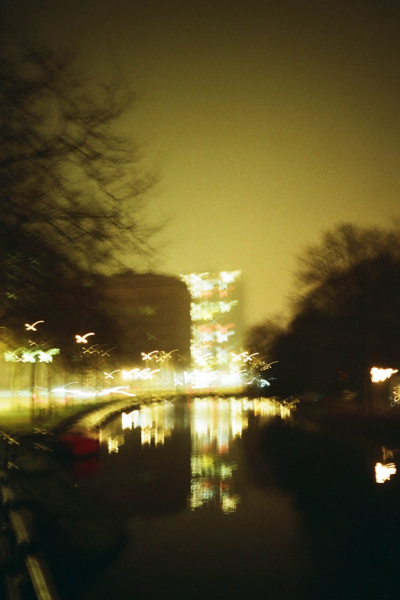 The image depicts a serene nighttime scene with a large body of water reflecting the lights around it. There's a bridge over the water that stretches across most of the frame. The sky above is dark, suggesting it's evening or nighttime. Streetlights and possibly other building lights are visible on the opposite shore, creating a gentle reflection in the water.